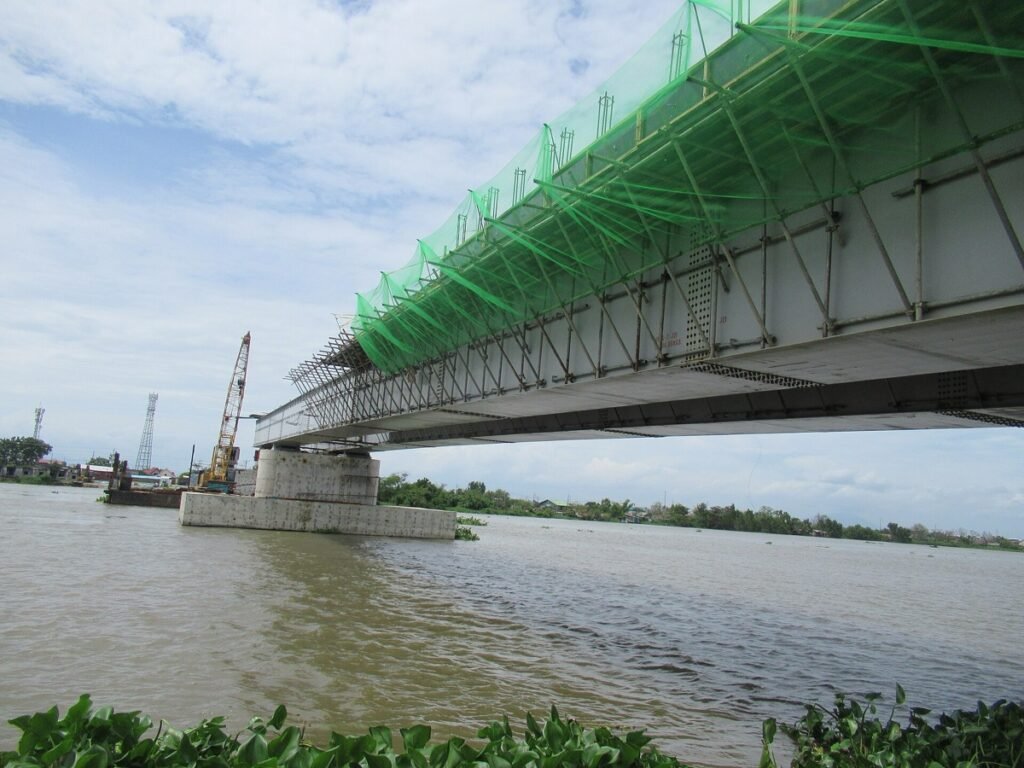 Pont en construction au-dessus d'une rivière, sous-face équipée d'échafaudages et de filets de protection.